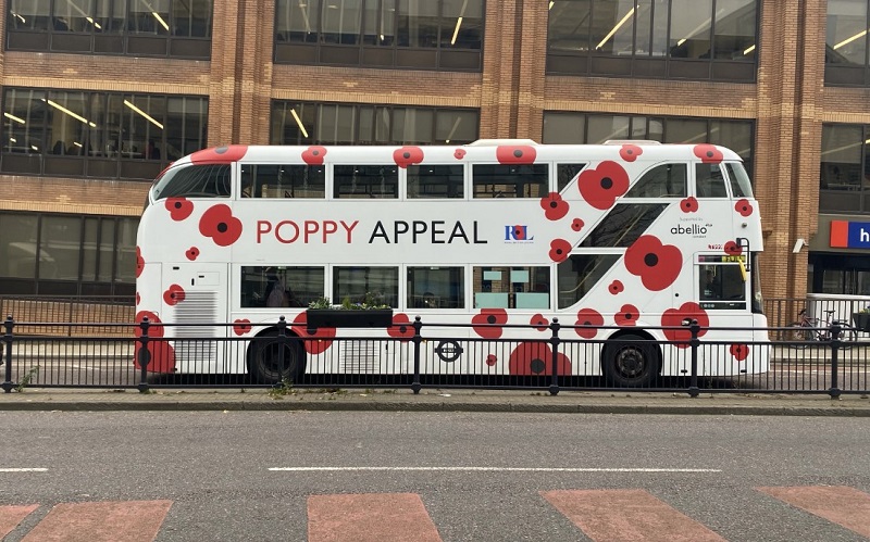 A bus with poppy appeal on the side driving down a street