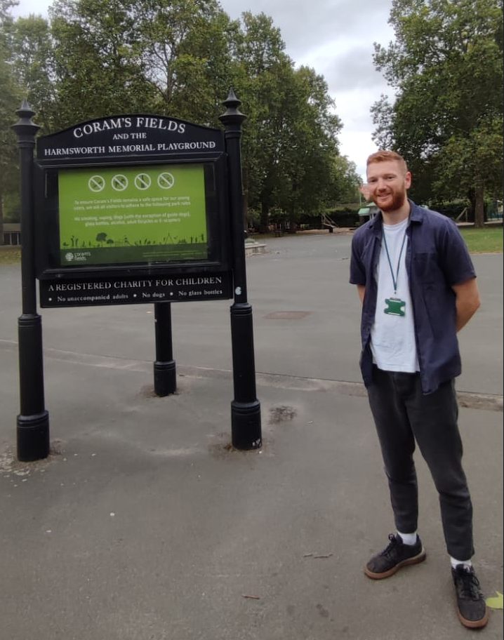 Ed May posing inside Coram's Fields in a purple shirt and dark-grey pants. The background includes large trees and a sign reading "Coram's Fields and the Harmsworth Memorial Playground".
