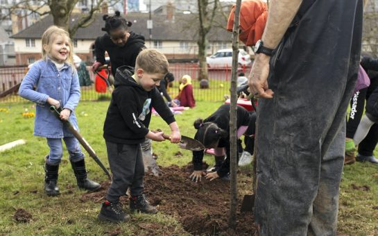 A group of children shoveling earth to bed in a newly planted apple tree, part of the Dove Gardens community garden initiative started by the residents themselves during lockdown, Bristol, March 2022