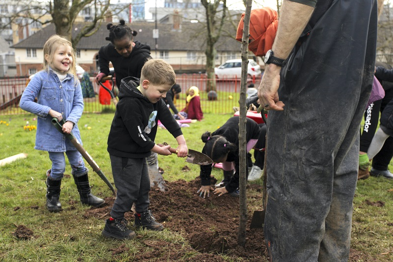 A group of children shoveling earth to bed in a newly planted apple tree, part of the Dove Gardens community garden initiative started by the residents themselves during lockdown, Bristol, March 2022