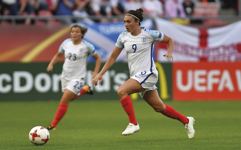 Image of two England women’s players in a match. They are in white kits with red socks