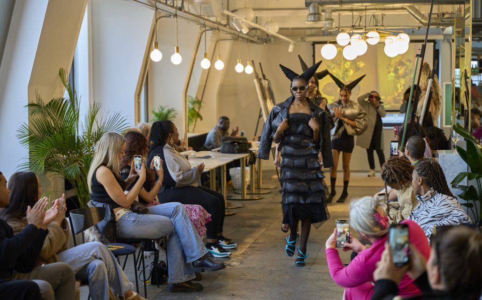 Photo of woman walking down improvised runway, wearing black dress and light blue shoes with hair styled in devil horns shape. People sitting by the side taking photos and videos on their phones
