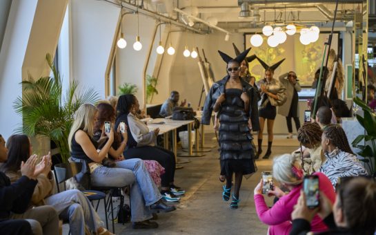 Photo of woman walking down improvised runway, wearing black dress and light blue shoes with hair styled in devil horns shape. People sitting by the side taking photos and videos on their phones