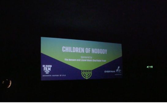 The screen at a cinema displaying a poster for the film Children of Nobody - white words saying "Children of Nobody" over a green background, and then purple background at the bottom of the screen with words saying "UK Jewish Film Festival 2023" and "Everyman"