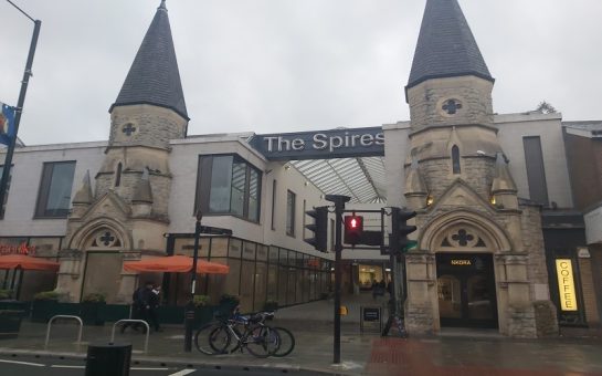 A photo of the outside of the Spires shopping centre on a grey day, showing the logo of the centre and the two spires for which it is named