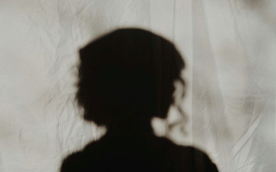 A photo of a woman's silhouette from the shoulders up. The background is a white/grey material and the woman is in shadow looking to the right of the image in side profile. She appears to have her hair up with curls falling above her shoulders.