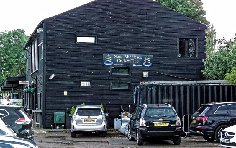 Image of North Middlesex CC clubhouse with visible banner on outside. Car parks with cars parked in is also visible