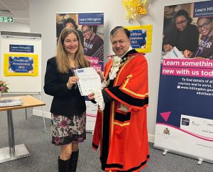 Oana-Maria-Vasile, adult learner accepting certificate from Mayor of Hillingdon in front of Adult Education posters in council offices.