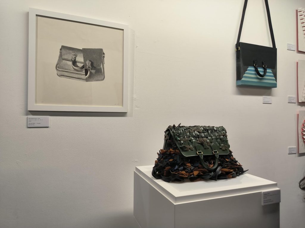 Image shows an exhibition display of different forms of crossbody bags: a framed pencil drawing hanging on the white wall, a remade green leather bag on a white block, and a plastic blue and black bag with a blue staircase painting at its centre, hanging on the wall.