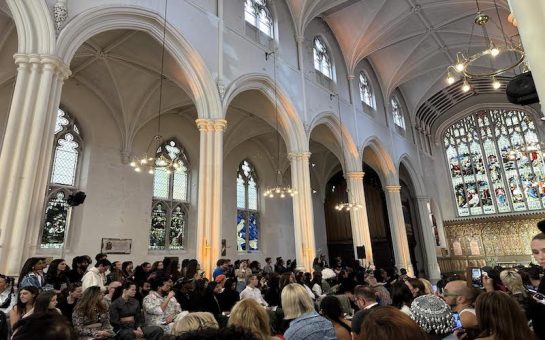 Image shows interior of a church with arches and a seated crowd lining the sides. A central runway has been staged, leading up to a large stained glass window.