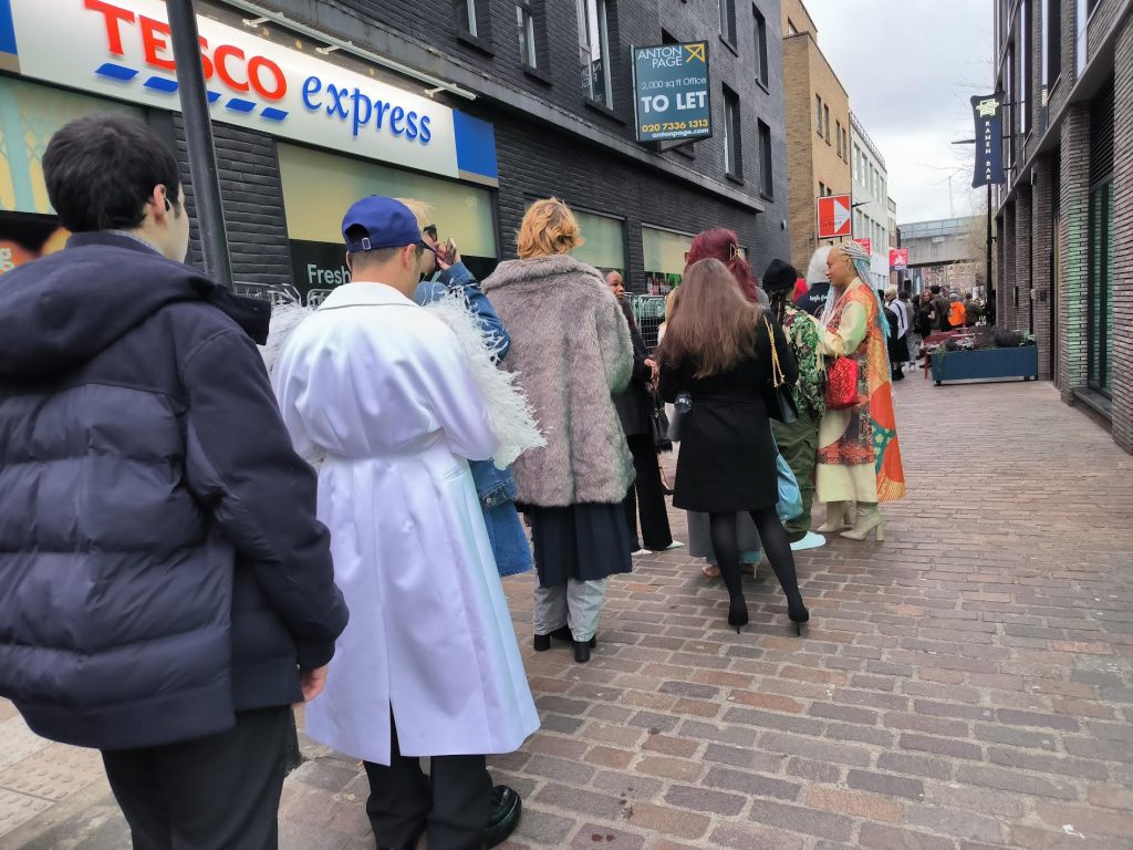 Image shows members of the public queuing for a fashion show, standing outside a brick building in the daytime.