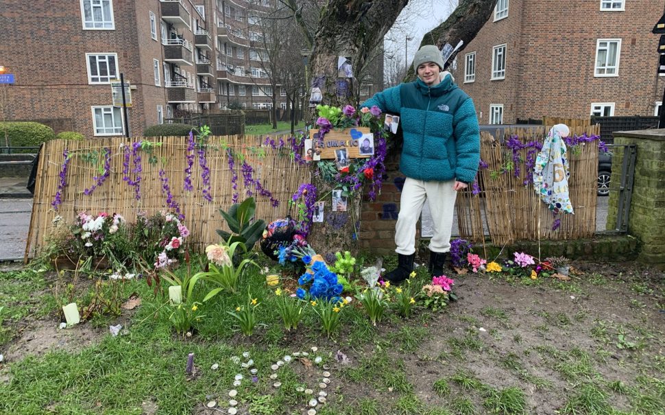 Young boy standing next to a tree in front of a fence. Tree and surrounding ground are covered in pictures and flowers of the deceased.