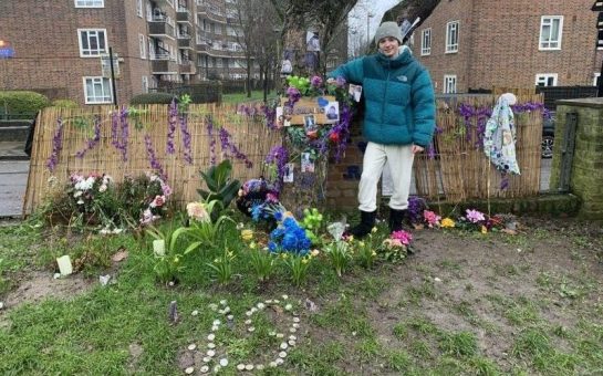 Young boy standing next to a tree in front of a fence. Tree and surrounding ground are covered in pictures and flowers of the deceased.