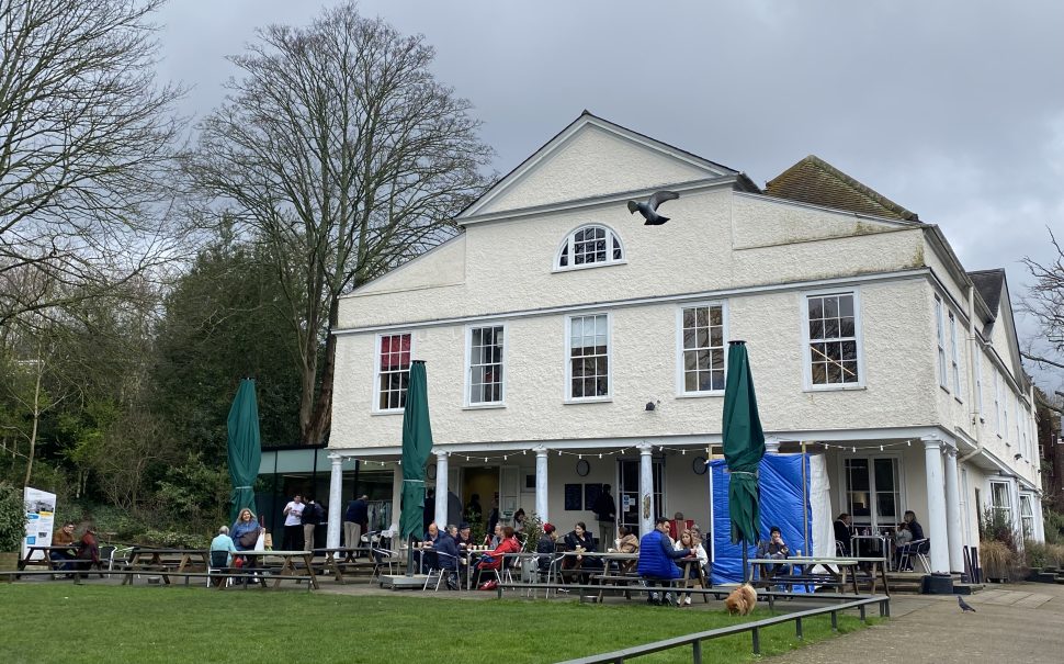 A large white house is verged by grass, and people sitting on picnic tables. Grey sky can be seen above.