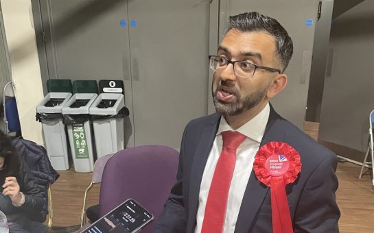 Krupesh Hirani talks to a reporter. He is looking to his left and wearing a black suit and white shirt with red tie and red Labour rousette