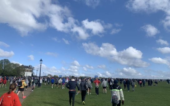 Runners and supporters walking towards the London Marathon starting line across a field on a sunny day.