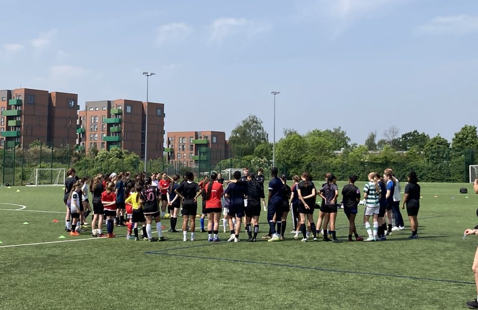 A group of around 60 women and girls on the pitch getting a briefing from coaches on how the trails would run. 