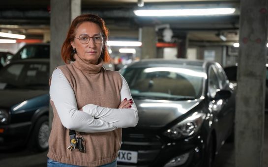 A woman stands in front of her car with her arms crossed looking angrily at the camera.