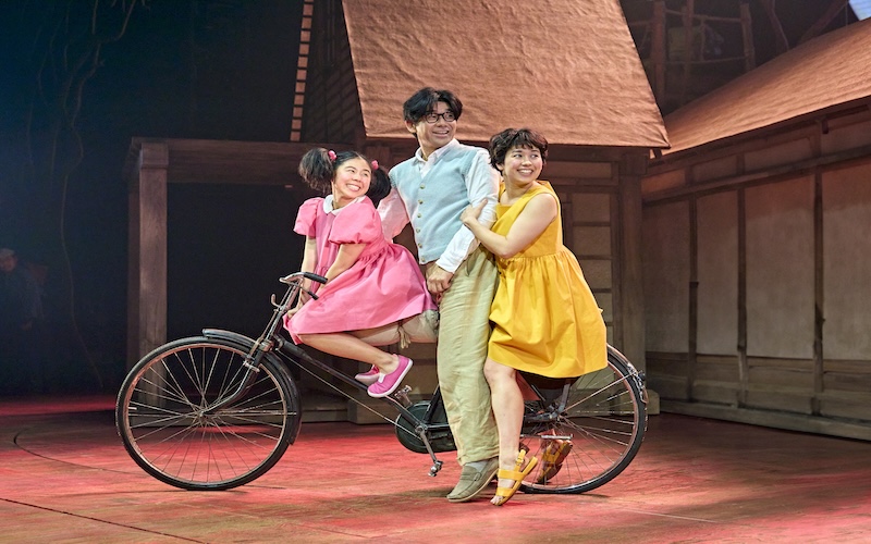 Mei (Victoria Chen), Tatsuo (Dai Tabuchi) and Satsuki (Ami Okumura Jones) all sitting on a bike together and looking back. (Picture credit: Manuel Harlan)