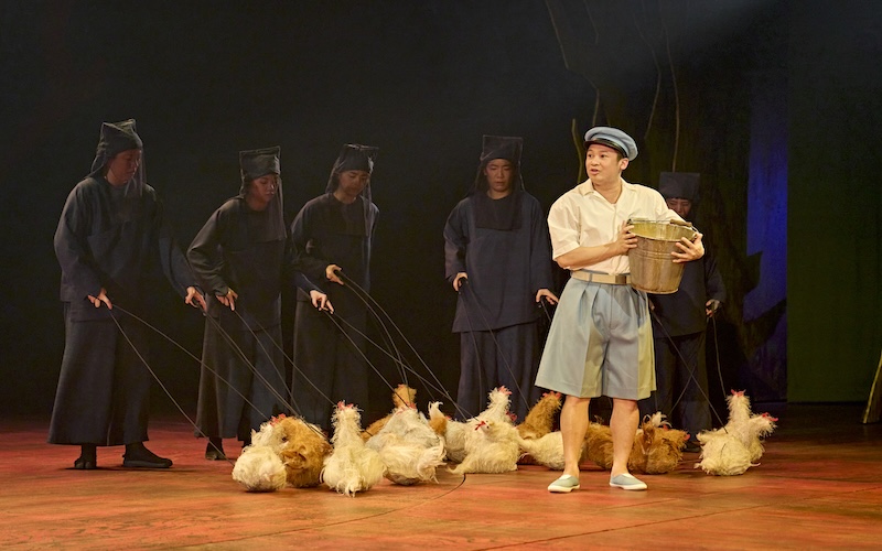 Kanta (Steven Nguyen) wearing a hat and carrying a basket with members of the company behind him with chicken puppets. (Picture credit: Manuel Harlan)