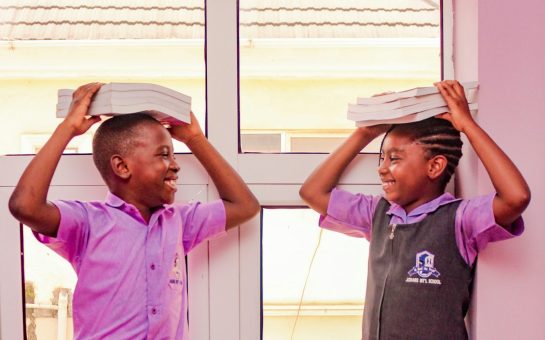 Two school children smiling at one another holding books on their head