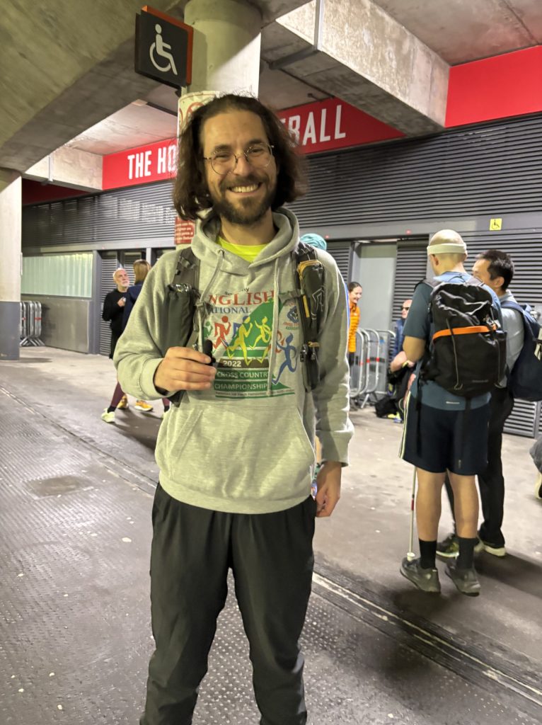 Simon Fitzmaurice, an Achilles UK guide runner, stood outside Emirates Stadium in a grey jumper with glasses and smiling.