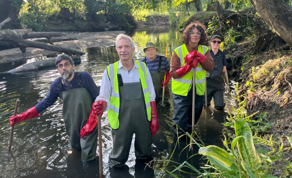 Two people in hi-vis jackets and three people in waterproof clothes behind them, all wearing red gloves, standing in a river, holding shovels and nets.