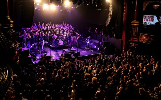 A zoomed-out concert view of a Soul Choir group singing on a stage, lit under purple lights, with the crowd clapping for them.