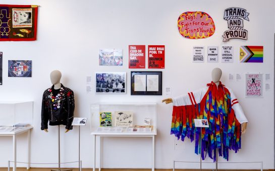 An LGBTQ+ exhibition in a museum in London, featuring two mannequins, tables with displays and LGBTQ posters on the wall.
