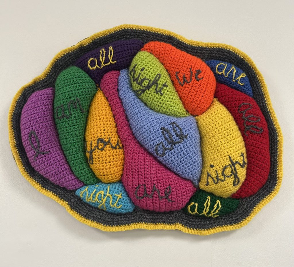 A multi-coloured crocheted and embroidered artwork by artist Leslie Wilson-Rutterford carrying the words, 'I Am All Right, You Are All Right, We Are All All Right', 2024. Displayed in Civic Action Hub, for 'From Kitchen Table to Gallery Wall' Art Exhibition. 