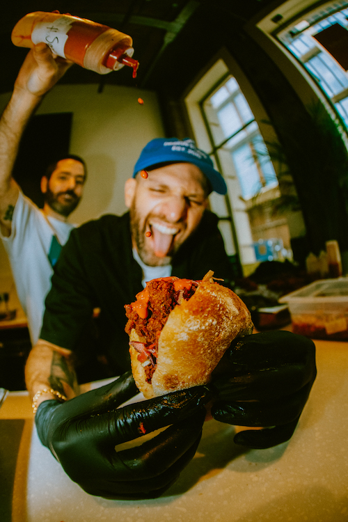 Sarnie Social founder Seif El-Sobky holds a fried chicken sandwich in a kitchen as a man pours hot sauce onto it.