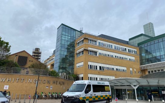 The front entrance to the Whittington Hospital. There is an ambulance parked outside the entrance to the hospital. There is a large sign that reads 'Whittington Health' on the side of the building. There are also Lime bikes parked outside.