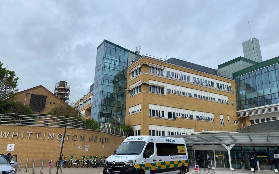 The front entrance to the Whittington Hospital. There is an ambulance parked outside the entrance to the hospital. There is a large sign that reads 'Whittington Health' on the side of the building. There are also Lime bikes parked outside.