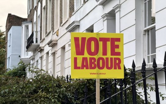 A bright yellow "Vote Labour" election campaign sign posted in front of white terraced houses along a street in Camden.