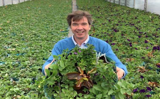 Miles Watson-Smyth holding a hanging basket inside a greenhouse filed with plants.