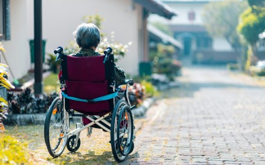 An elderly woman in a wheelchair sitting alone on the side of a pavement.
