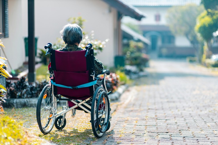 An elderly woman in a wheelchair sitting alone on the side of a pavement.