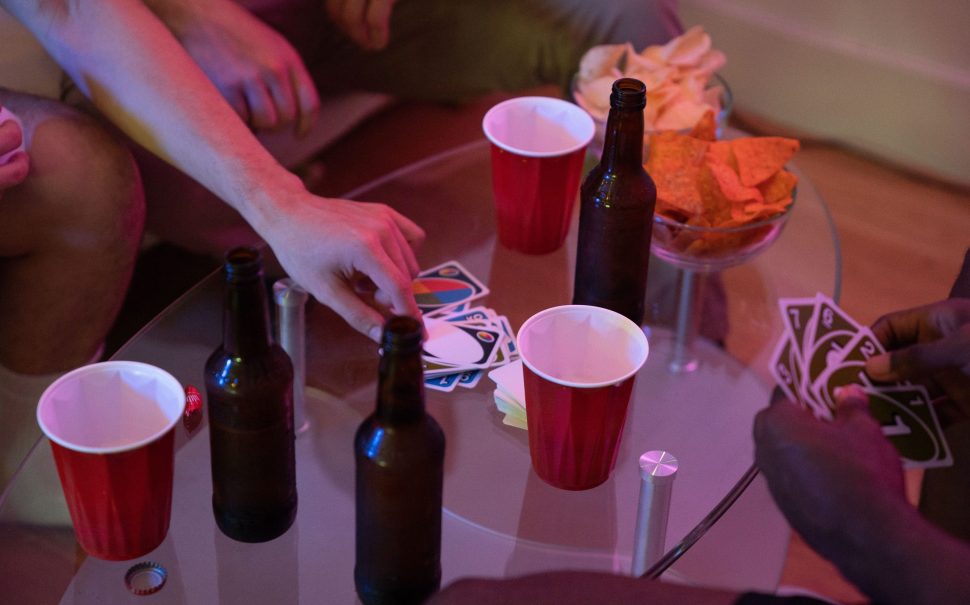 Group of people playing a card game around a glass coffee table with beer bottles, plastic cups, and bowls of chips